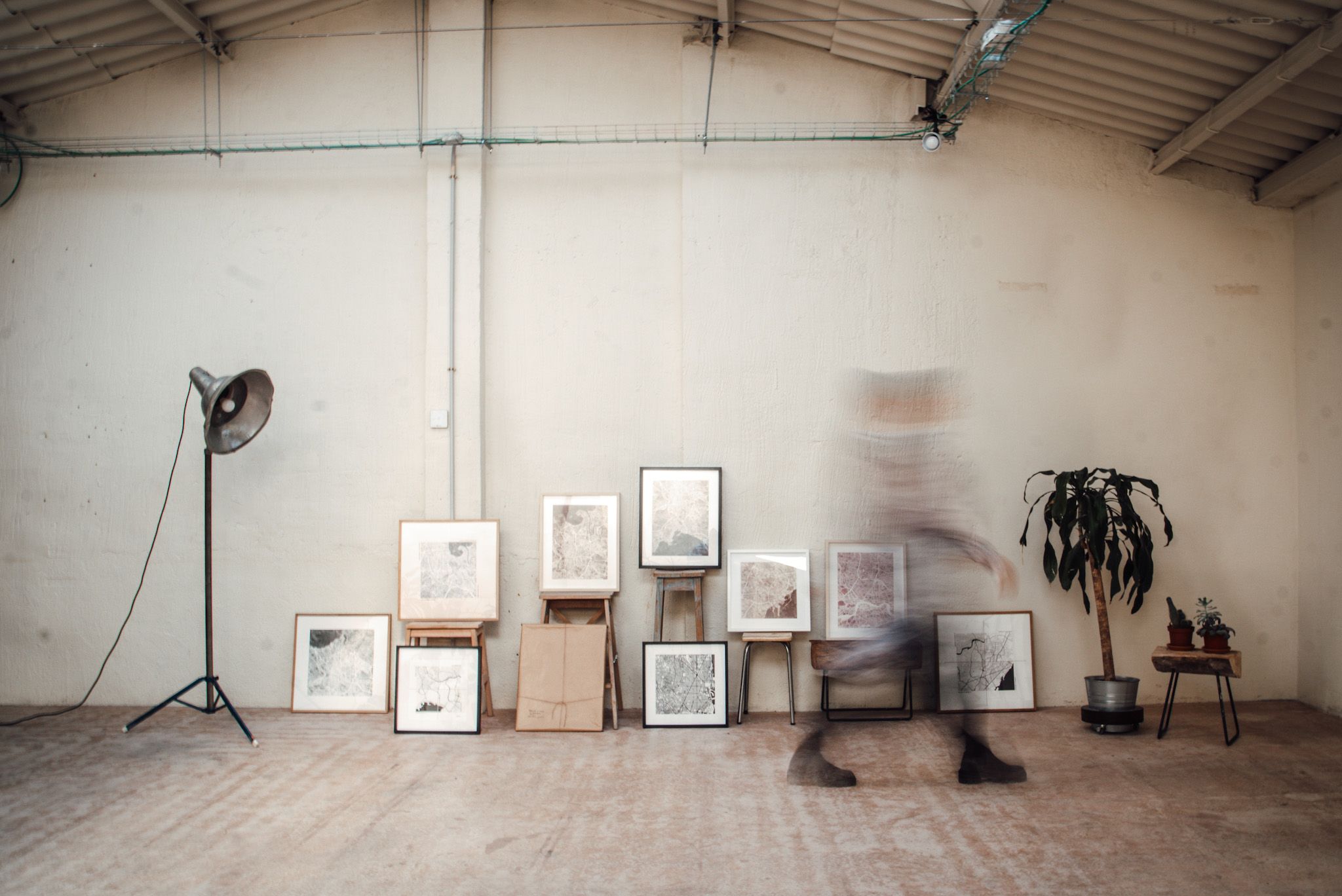 Imagen minimalista de una nave con cuadros, una lámpara estilo industrial, unas plantas y una mujer andando difuminada